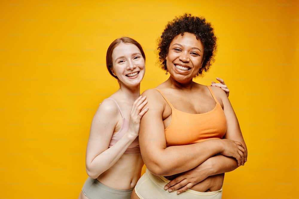 Retrato de cintura para arriba de dos mujeres jóvenes sonrientes de piel oscura y piel clara abrazándose mientras están de pie contra un fondo amarillo vibrante en ropa interior
