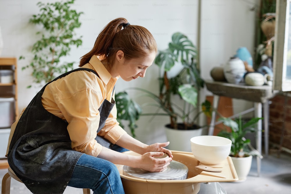 Side view portrait of young woman using pottery wheel and creating handmade ceramics in cozy studio