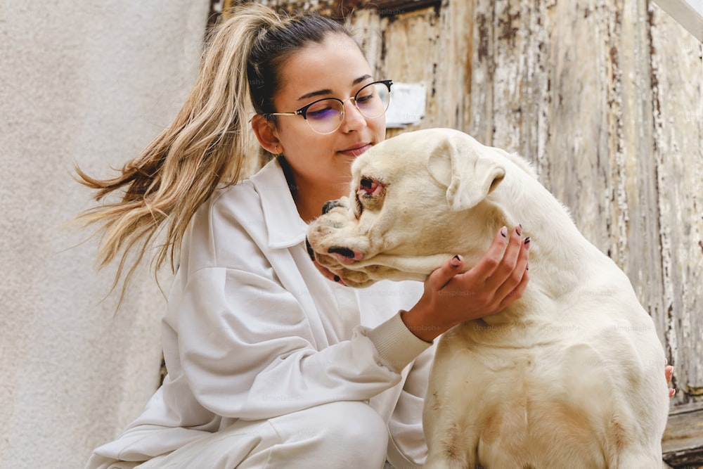 白い犬と遊んで世話をしている日、家の前の階段に座っている若い白人女性 – 友情とペット愛のコンセプト実在の人々の明るい写真の正面図