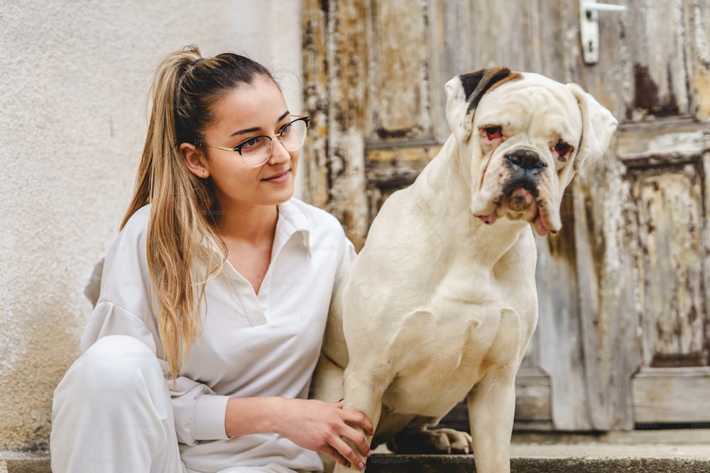 Young caucasian woman sitting on the stairs in front of her home in day with white dog playing with him and taking care of - friendship and pet love concept real people bright photo front view
