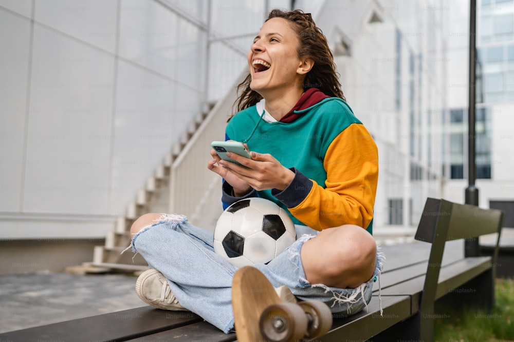 One young caucasian woman female sitting on the bench in front of the building or at stadium with soccer ball waiting for the football game using mobile phone happy smile real people copy space