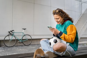 One young caucasian woman female sitting on the bench in front of the building or at stadium with soccer ball waiting for the football game using mobile phone real people copy space