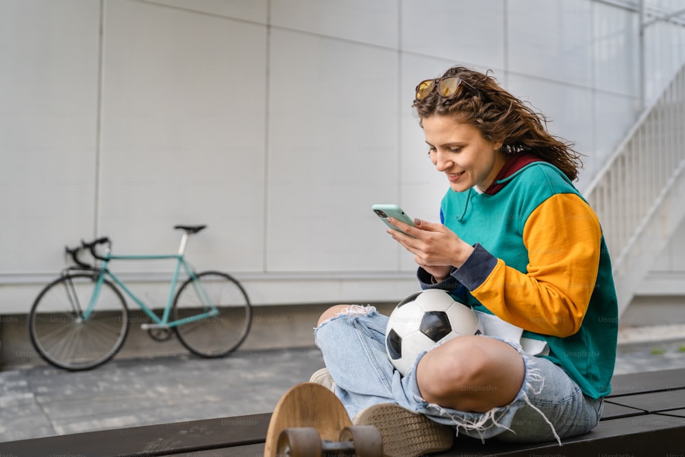 Una joven mujer caucásica sentada en el banco frente al edificio o en el estadio con balón de fútbol esperando el partido de fútbol usando el teléfono móvil personas reales copian el espacio