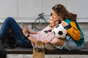 Couple woman and young caucasian man modern happy adult male and female boyfriend and girlfriend smile sitting in front of stadium holding soccer ball outdoor in day football game concept copy space