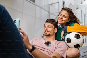 Pareja mujer y joven hombre caucásico moderno feliz adulto masculino y femenino novio y novia sonríe sentado frente al estadio sosteniendo balón de fútbol al aire libre en el concepto de juego de fútbol de día copiar espacio
