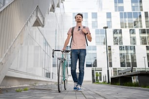 one man young adult caucasian male walking by the modern building with his bicycle in day full length front view copy space male student in front of the university casual lifestyle concept
