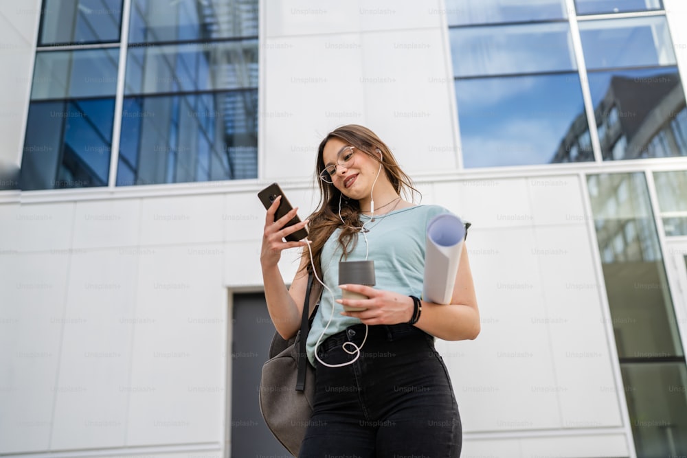 Uma mulher jovem adulta caucasiana estudante do sexo feminino em pé na frente da universidade com papel e xícara de café ao ar livre no dia brilhante usando o telefone celular esperando sorriso feliz pessoas reais copiar espaço