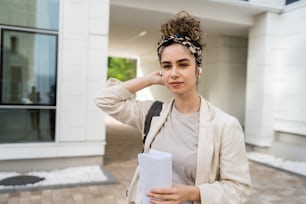 Eine junge Frau, eine Geschäftsfrau oder eine Studentin, die Dokumente vor einem modernen Gebäude an einer Universität oder einem Unternehmen an hellem Tag überprüft, echte Menschen, die allein stehen, Papier halten