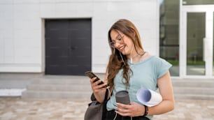 One woman young adult caucasian female student standing in front of university with paper and cup of coffee outdoor in bright day using mobile phone waiting happy smile real people copy space