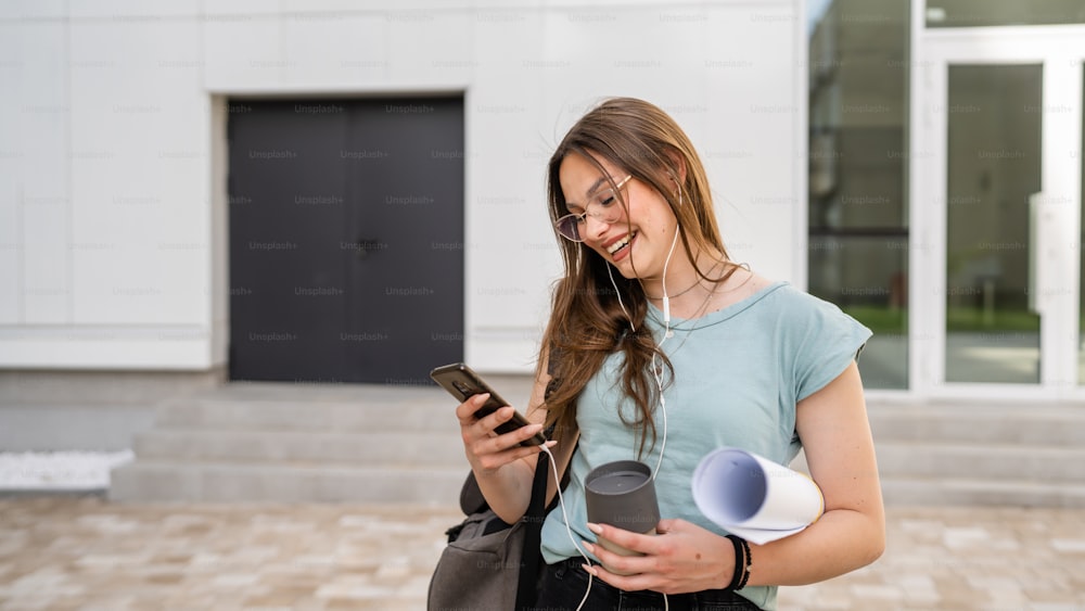 Eine junge erwachsene kaukasische Studentin steht vor der Universität mit Papier und einer Tasse Kaffee im Freien an hellem Tag mit Handy und wartet glücklich lächeln echte Menschen kopieren Raum