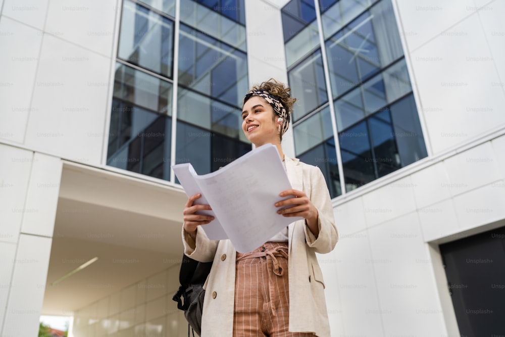 One young woman female businessman or student checking documents in front of modern building at university or company in bright day real people copy space standing alone hold paper happy smile