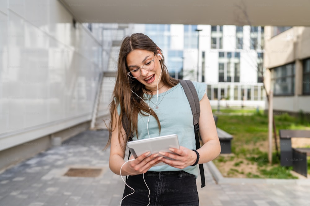 Uma mulher jovem adulta caucasiana estudante do sexo feminino em pé na frente do edifício da universidade ao ar livre no dia brilhante usando tablet digital esperando sorriso feliz pessoas reais copiar espaço