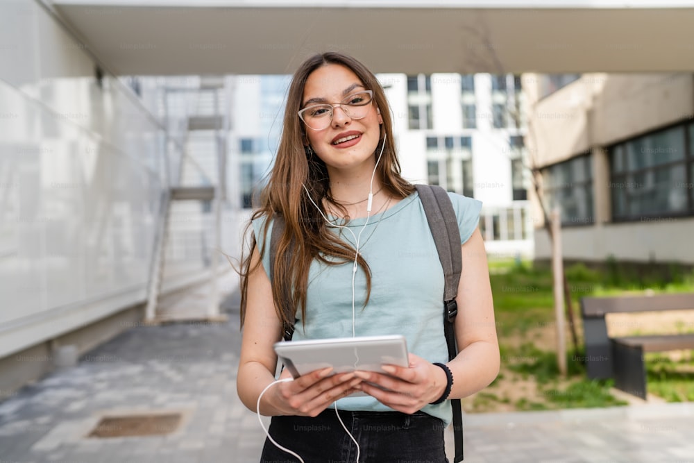 Eine junge erwachsene kaukasische Studentin steht vor dem Universitätsgebäude im Freien an hellem Tag mit digitalem Tablet und wartet glücklich lächeln echte Menschen kopieren Raum