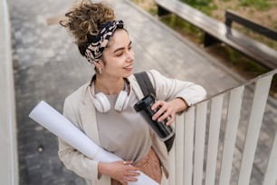 Eine Frau junge erwachsene kaukasische Studentin mit Rucksack steht oder geht vor dem Universitätsgebäude mit Papier und Tasse Kaffee im Freien in hellem Tag glückliches Lächeln echte Menschen kopieren Raum