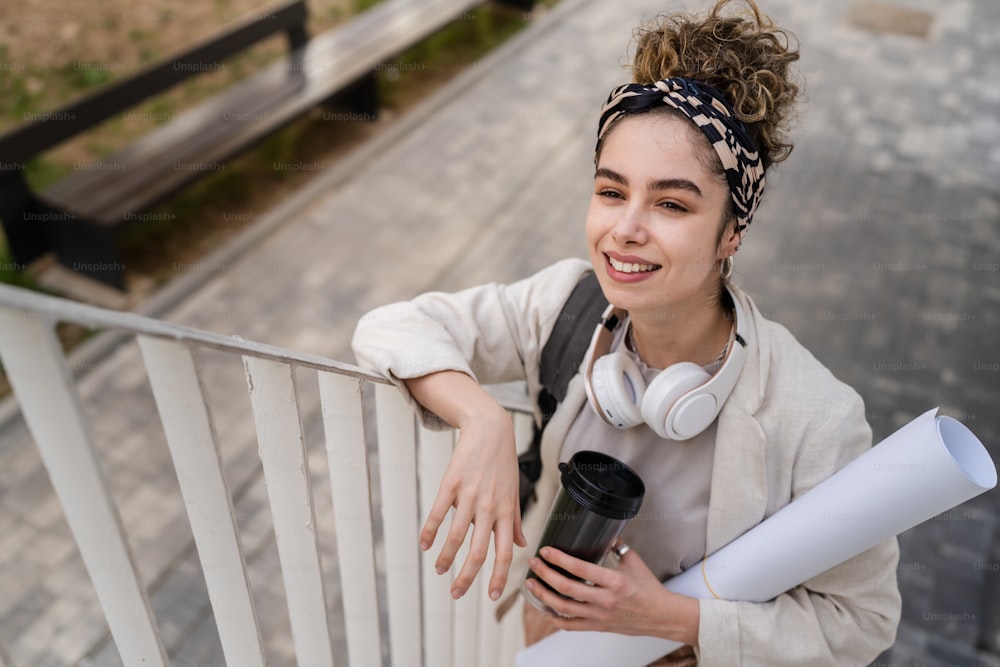 Uma mulher jovem adulta caucasiana estudante do sexo feminino com mochila em pé ou andando em frente ao prédio da universidade com papel e xícara de café ao ar livre em dia brilhante sorriso feliz pessoas reais copiar espaço