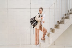 One woman young adult caucasian female student with backpack standing or walking in front of university building with paper and cup of coffee outdoor in bright day happy smile real people copy space