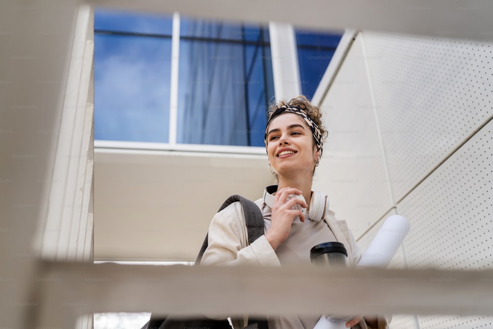 One woman young adult caucasian female student with backpack standing or walking in front of university building with paper and cup of coffee outdoor in bright day happy smile real people copy space