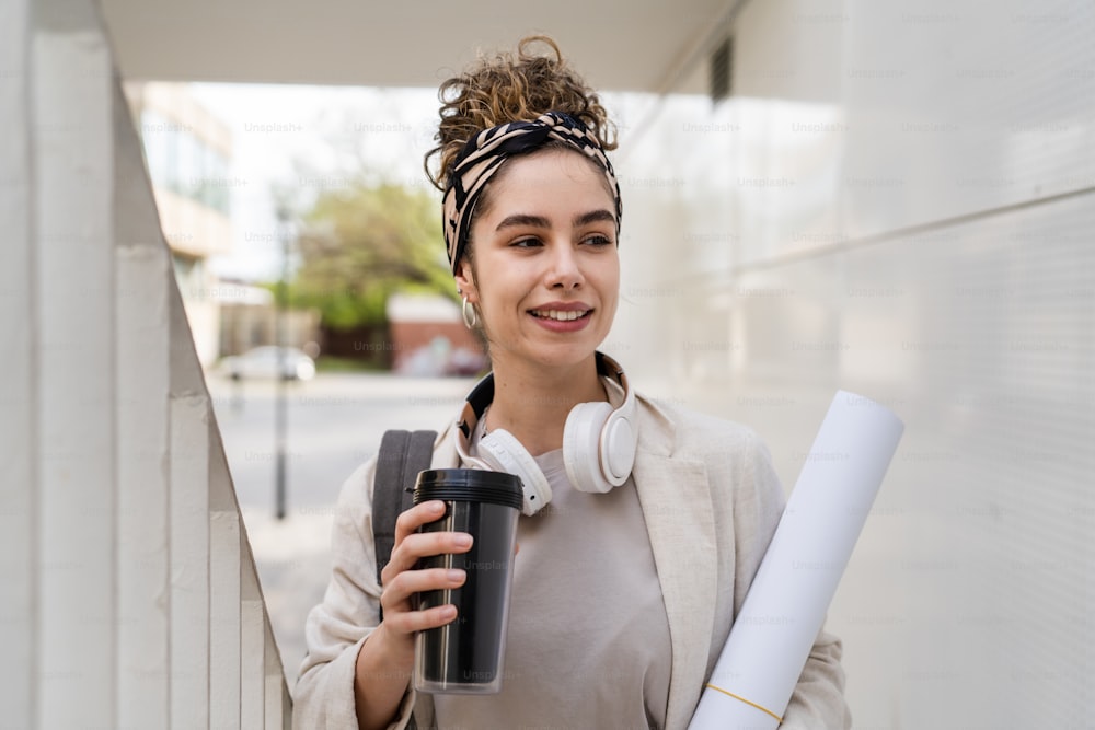 One woman young adult caucasian female student with backpack standing or walking in front of university building with paper and cup of coffee outdoor in bright day happy smile real people copy space