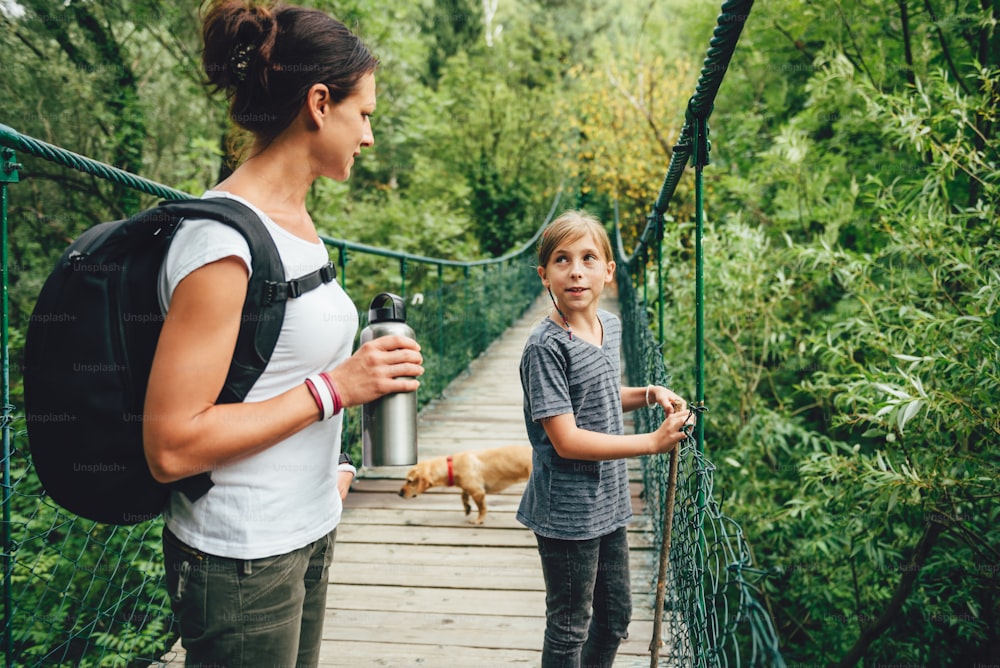 Mother and daughter standing on wooden suspension bridge with small yellow dog and talking