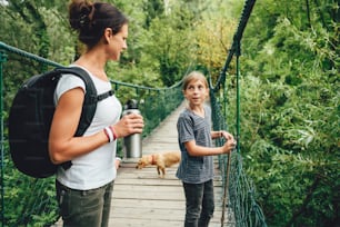 Mother and daughter standing on wooden suspension bridge with small yellow dog and talking