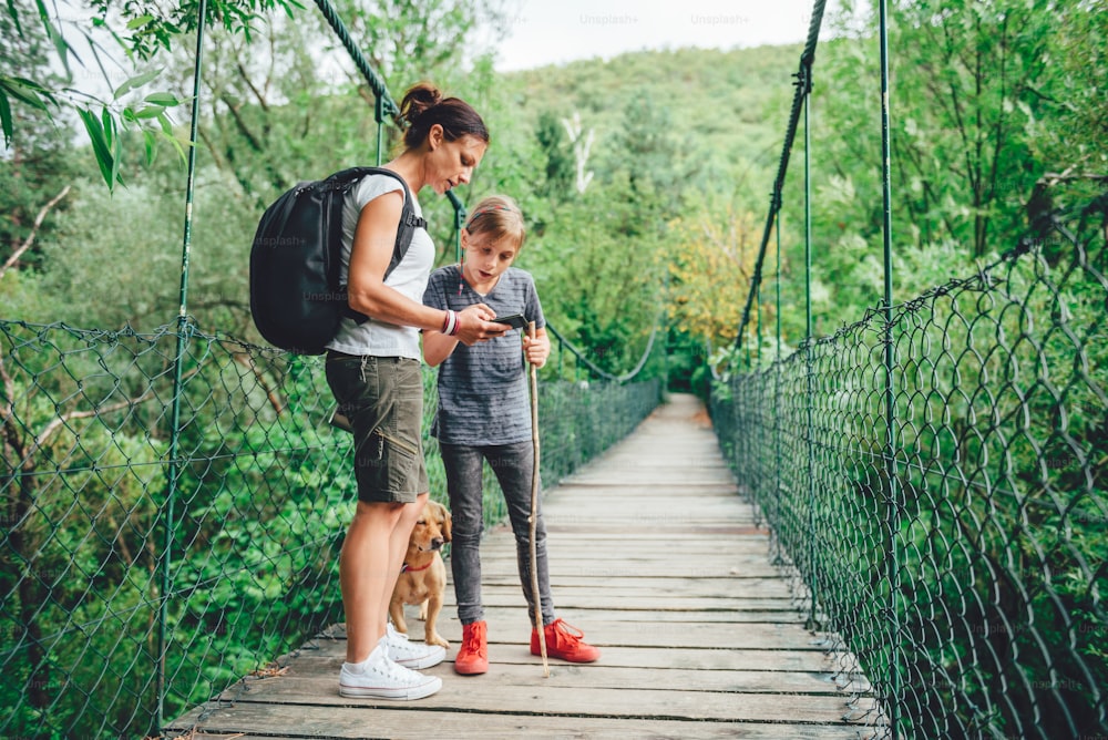 Mutter und Tochter stehen auf einer hölzernen Hängebrücke mit einem kleinen gelben Hund im Wald und benutzen ein Smartphone, um zu navigieren