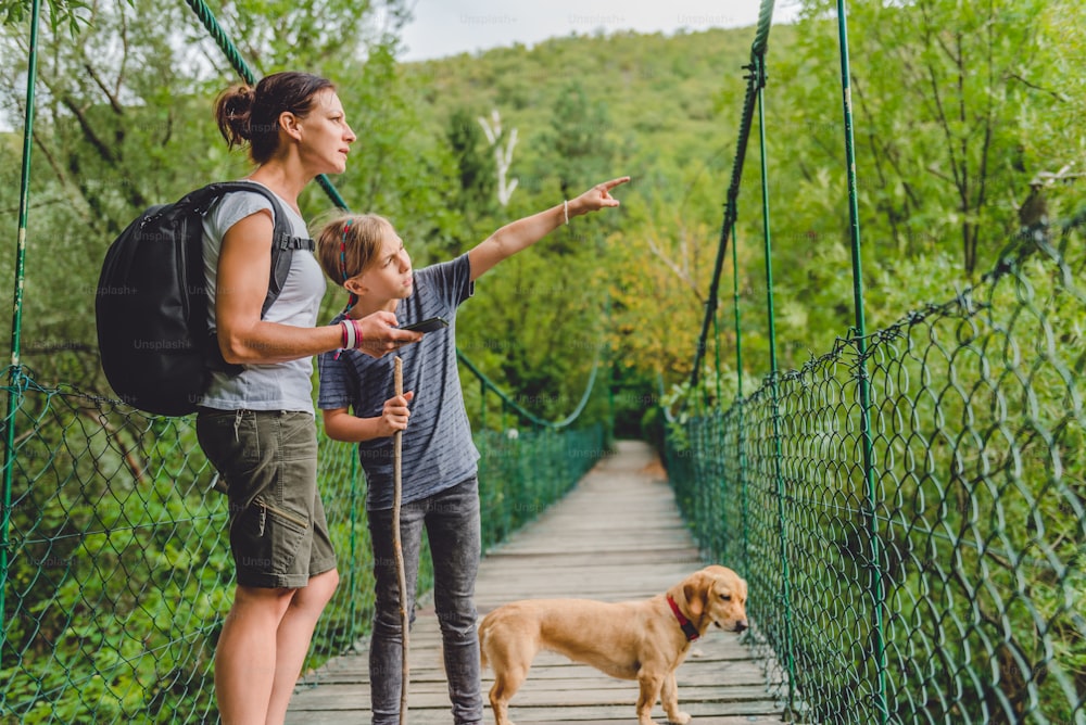 Mother and daughter standing on wooden suspension bridge with small yellow dog in the forest and using smart phone to navigate