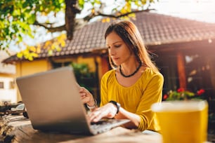 Women wearing yellow shirt sitting in the backyard patio, shopping online on laptop and using credit card