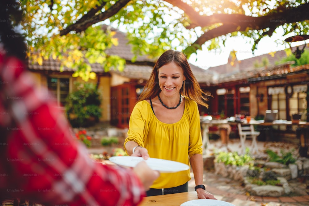 Woman with long brown hair wearing yellow shirt placing dishes on the table at backyard patio