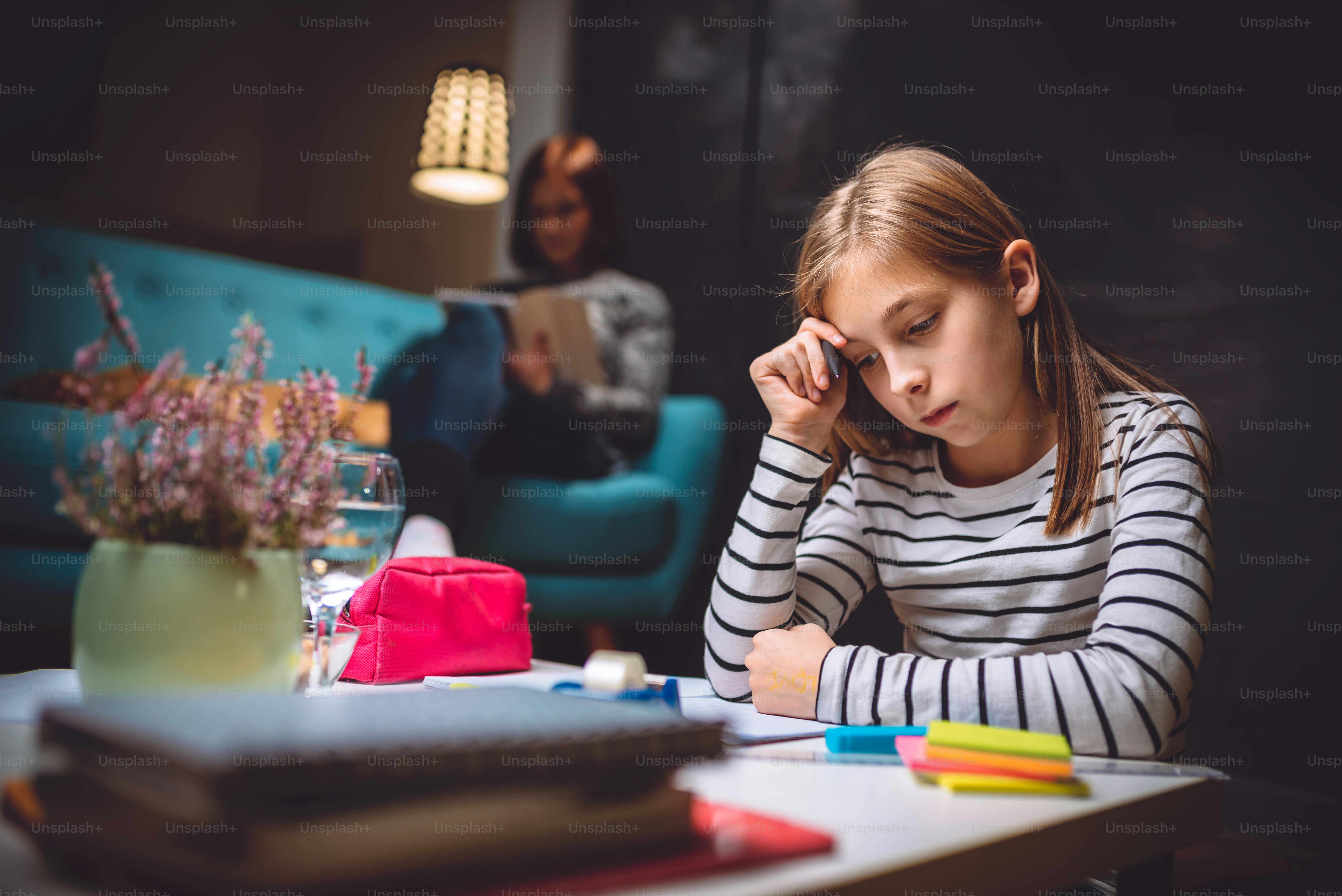 Girl sitting at coffee table in living room with her mother in the background and doing homework late at night