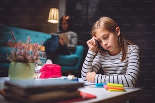 Girl sitting at coffee table in living room with her mother in the background and doing homework late at night