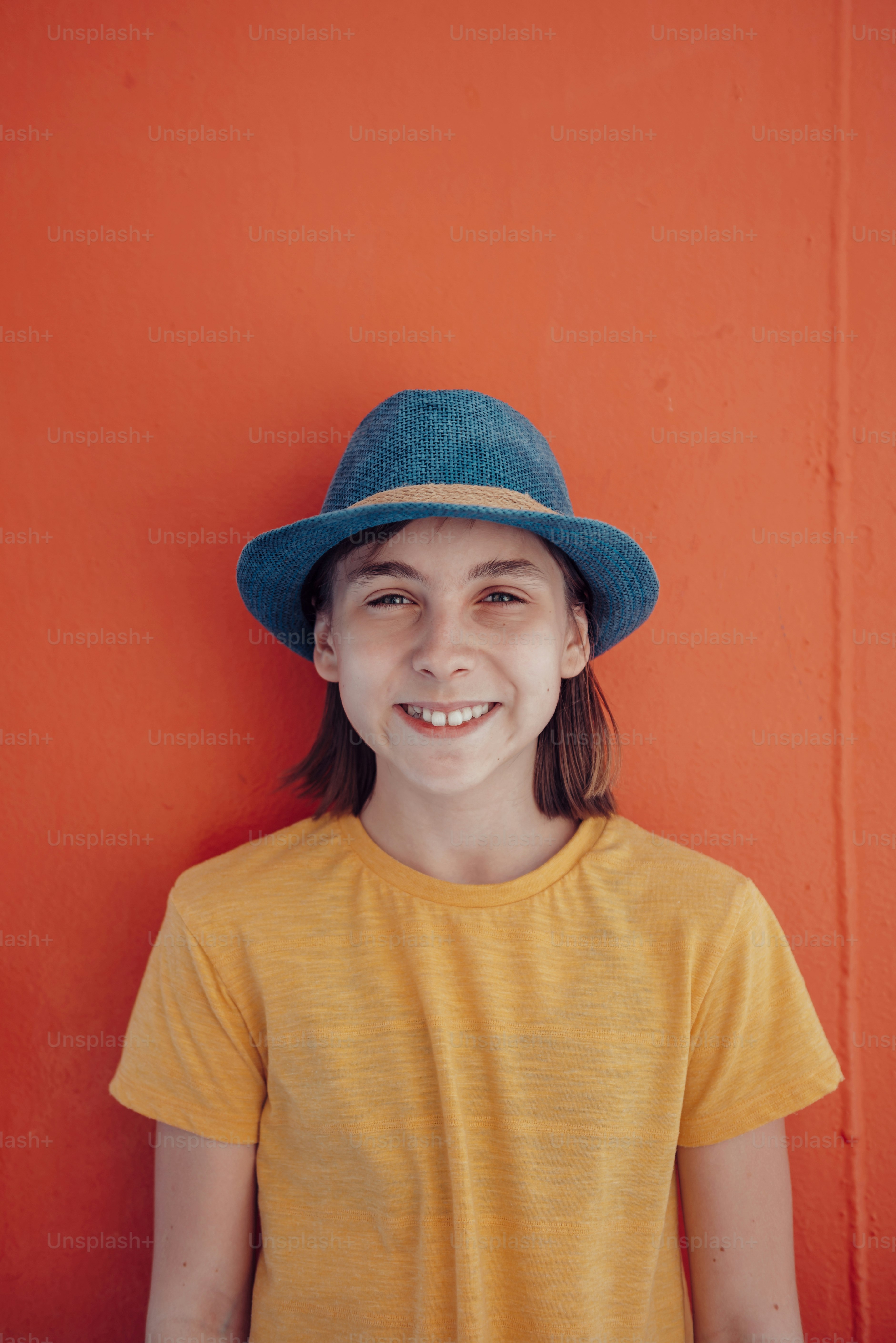 Retrato da menina que veste o chapéu do sol azul em pé contra a parede vermelha e sorrindo