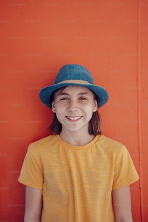 Portrait of girl wearing blue sun hat standing against red wall and smiling