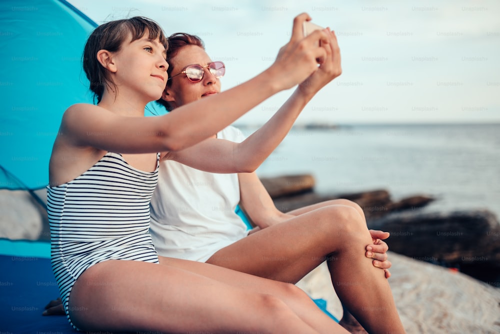 Filha e mãe tirando selfie com smartphone enquanto sentadas dentro da tenda de praia azul à beira-mar