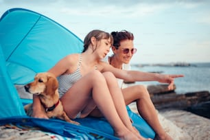母親と一緒に青いビーチテントの中に座り、小さな茶色の犬を抱きしめながら海を指さす女の子