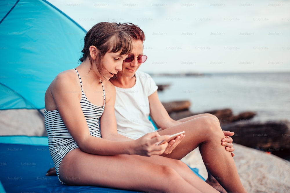 Madre e figlia che usano lo smartphone mentre sono sedute all'interno della tenda da spiaggia blu in riva al mare