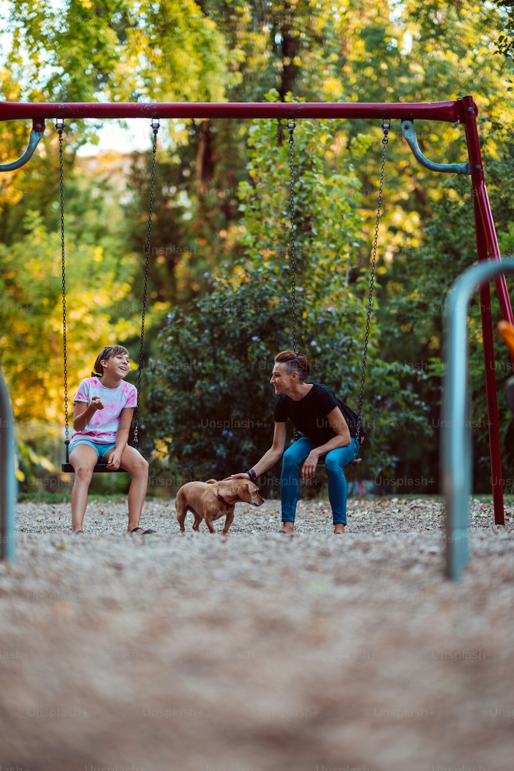 Madre e hija con un pequeño perro marrón sentados al aire libre en el columpio en el patio de recreo y hablando
