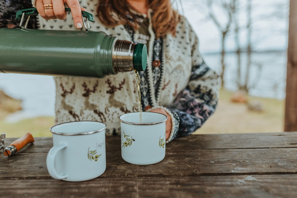 a woman pouring something into two mugs on a table