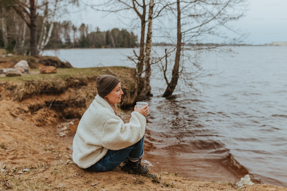 a woman sitting on the shore of a lake drinking a cup of coffee