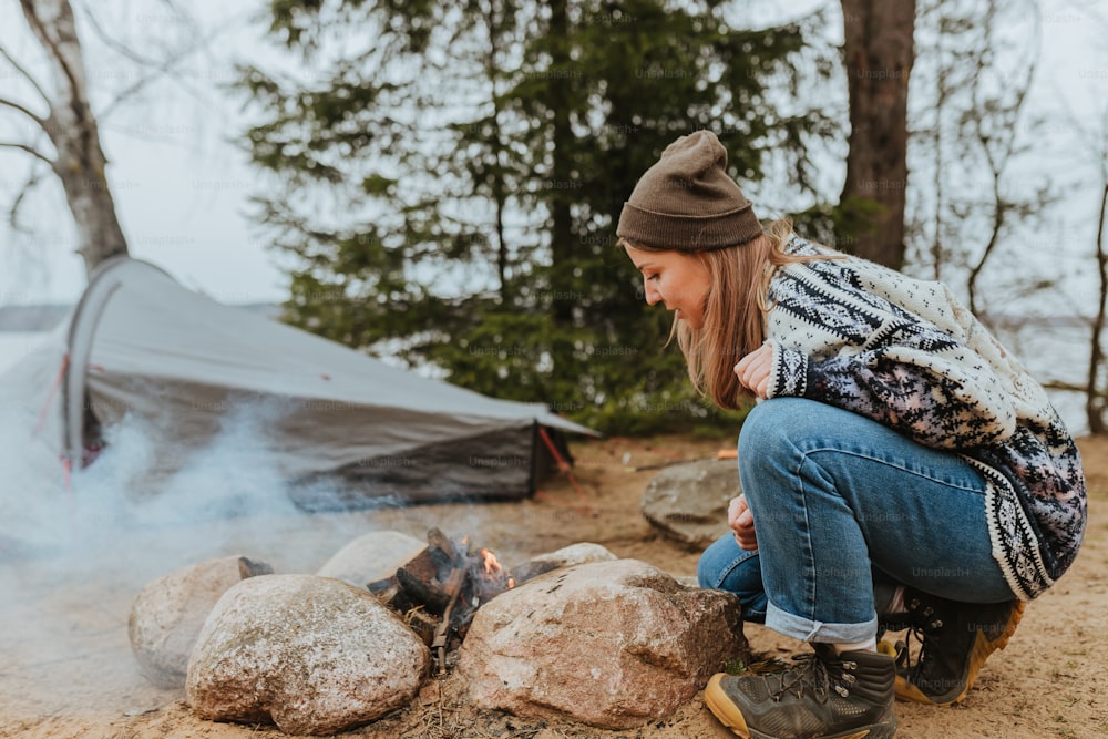 a woman squatting next to a campfire with a tent in the background