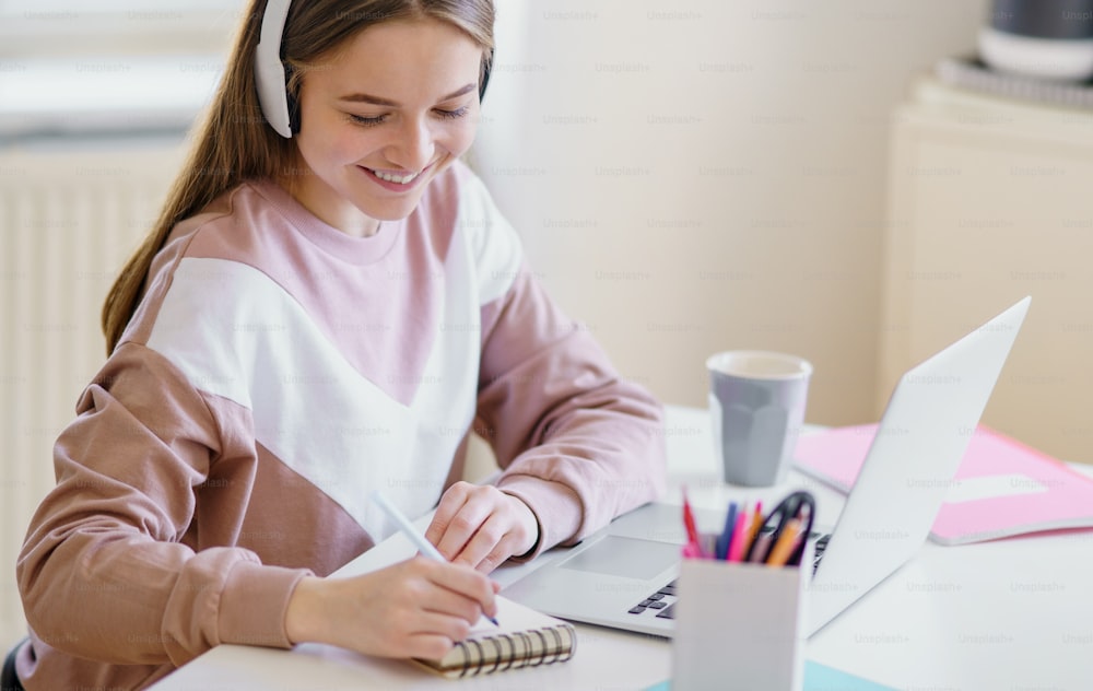 Junge glückliche College-Studentin, die zu Hause am Tisch sitzt und beim Lernen Laptop und Kopfhörer benutzt.