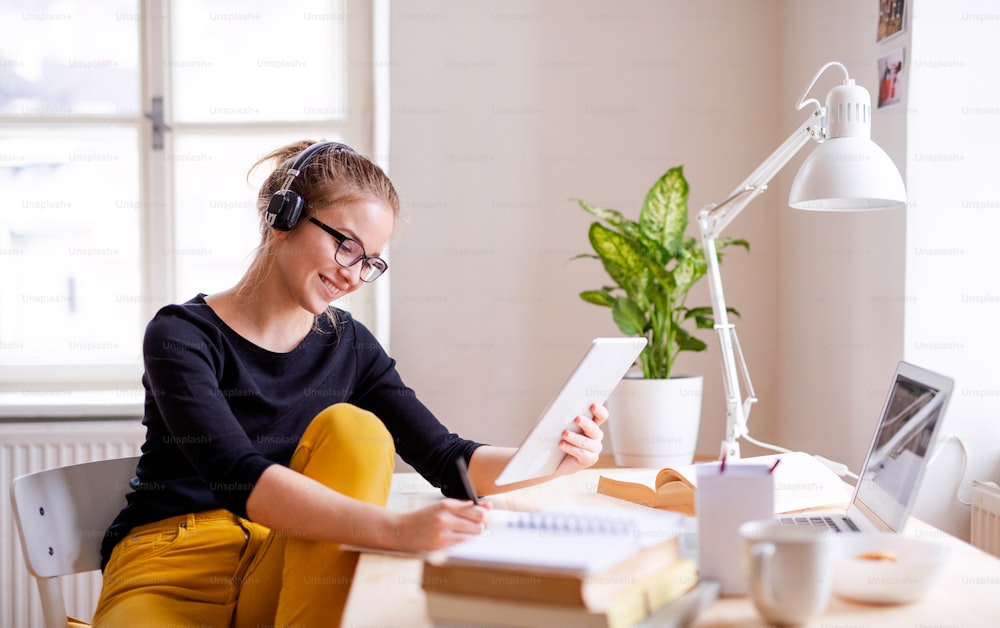 Une jeune étudiante heureuse assise à la table à la maison, utilisant une tablette et des écouteurs pendant ses études.