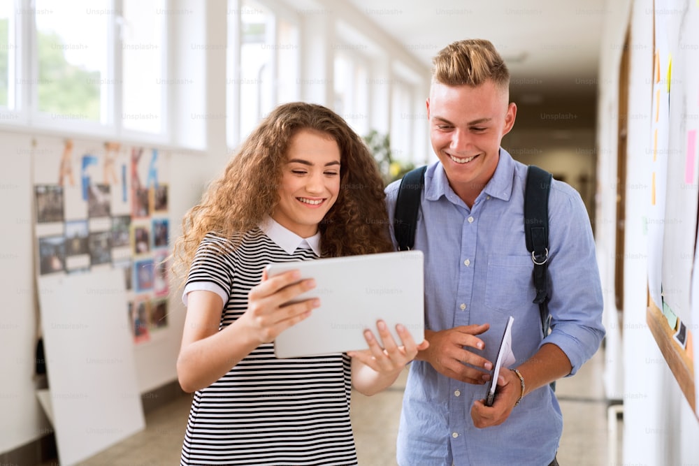 Attractive teenage student couple in high school hall with tablet, taking selfie.