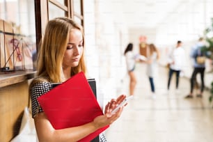 Attractive teenage girl with smartphone and notebooksin high school hall.