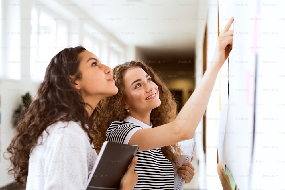 Two attractive teenage girls in high school hall during break reading something on notice board.