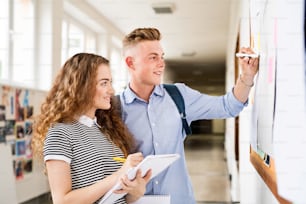 Atractiva pareja de estudiantes adolescentes en el pasillo de la escuela secundaria escribiendo algo en el tablón de anuncios.