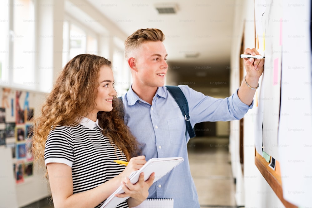 Attractive teenage student couple in high school hall writing something on notice board.