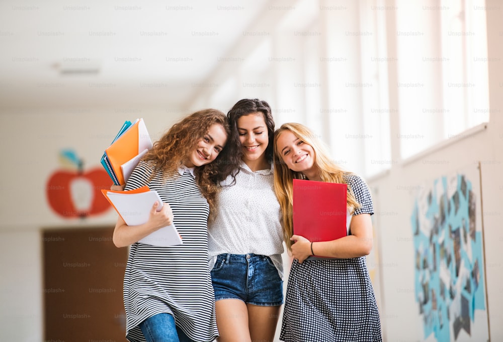 Group of three attractive teenage girls in high school hall during break, hugging and smiling.