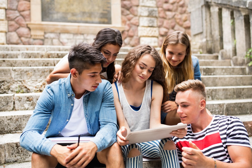 Grupo de estudiantes adolescentes atractivos sentados en escalones de piedra frente a la universidad sosteniendo una tableta, leyendo o mirando algo.