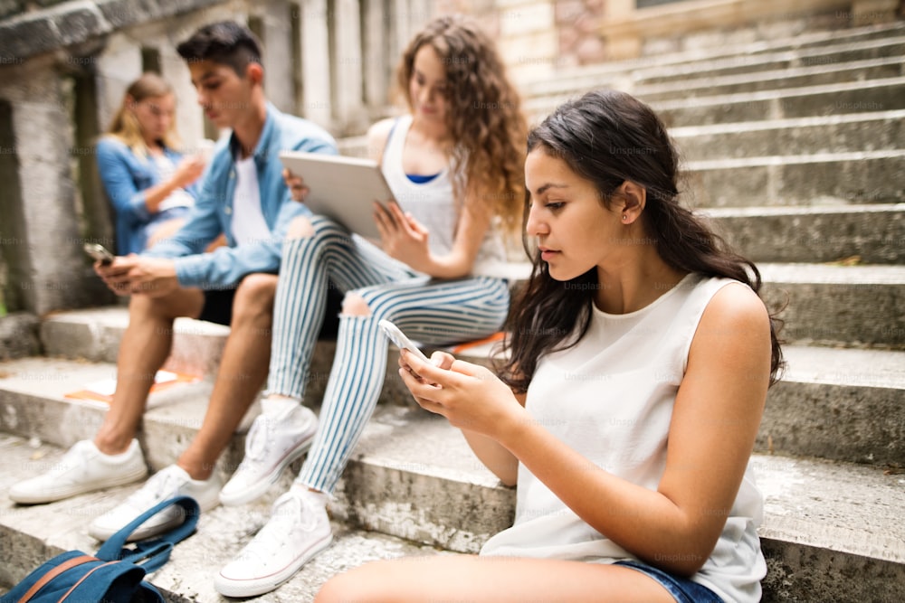 Attractive teenage students sitting on stone steps in front of university holding smart phones and tablet, reading or watching something.