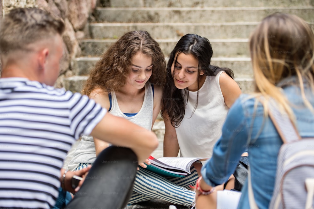 Group of four attractive teenage students sitting on stone steps in front of university studying.